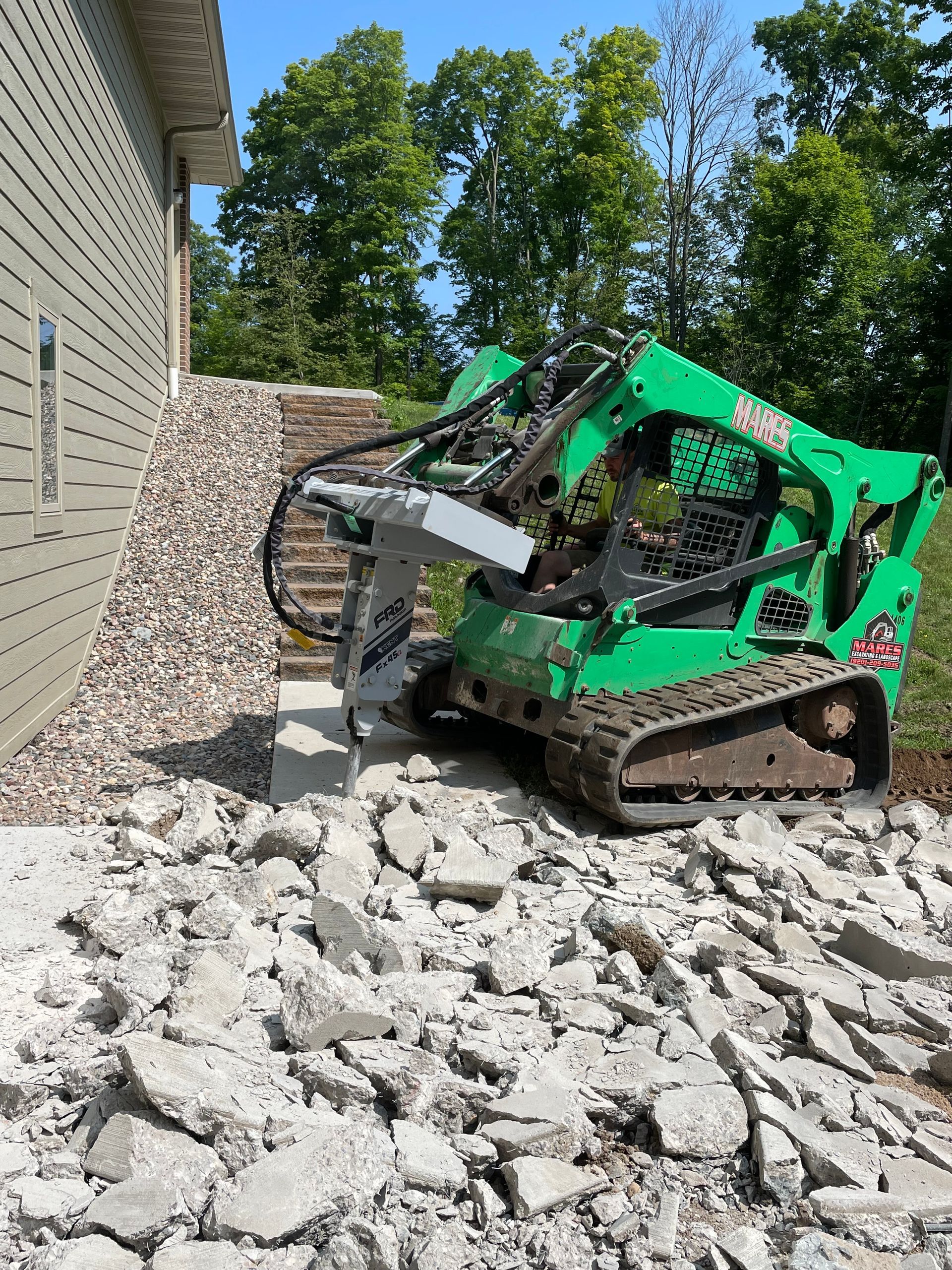 A green bulldozer is breaking up concrete in front of a house.