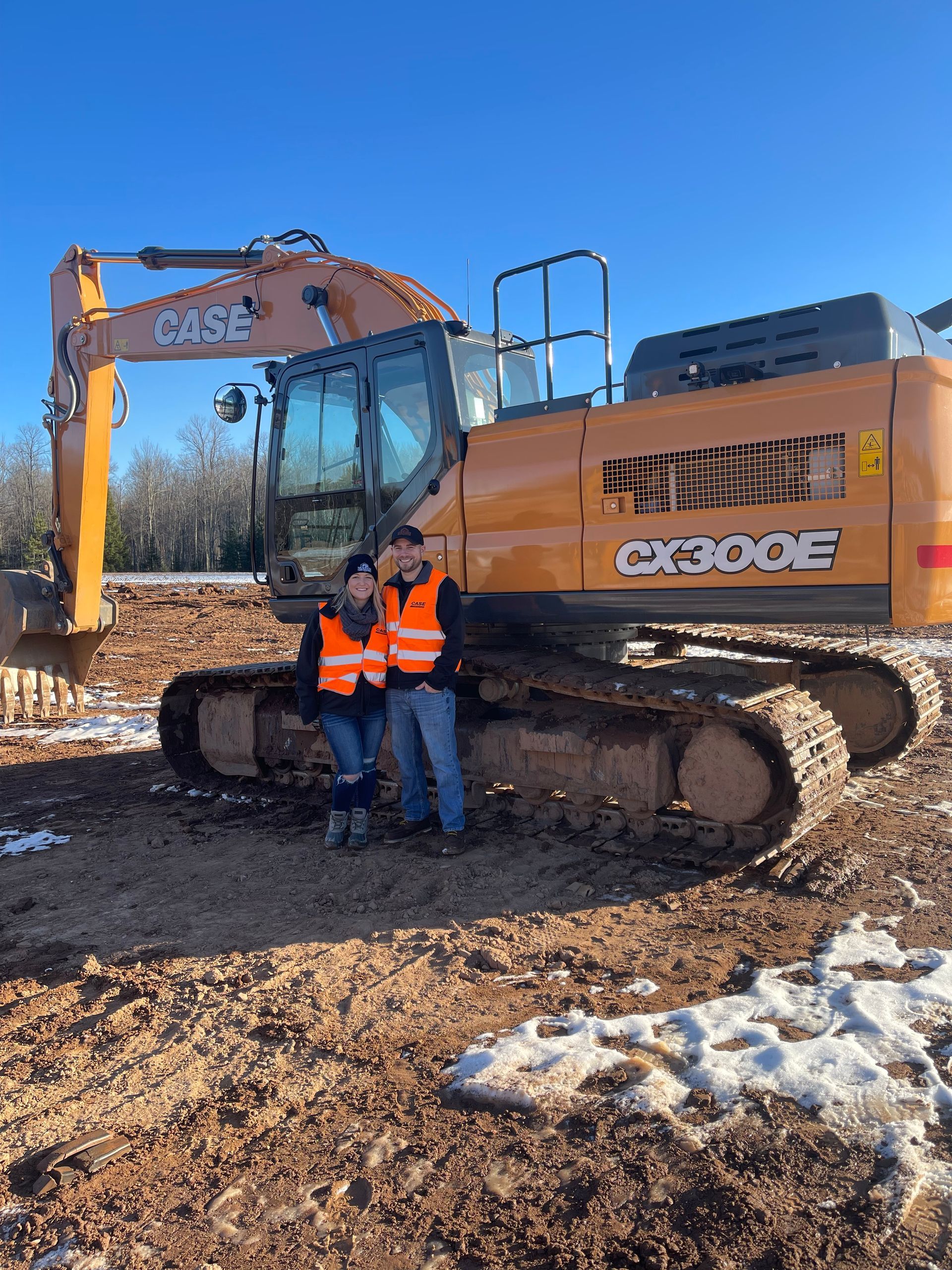 Two men are standing in front of an excavator in a dirt field.