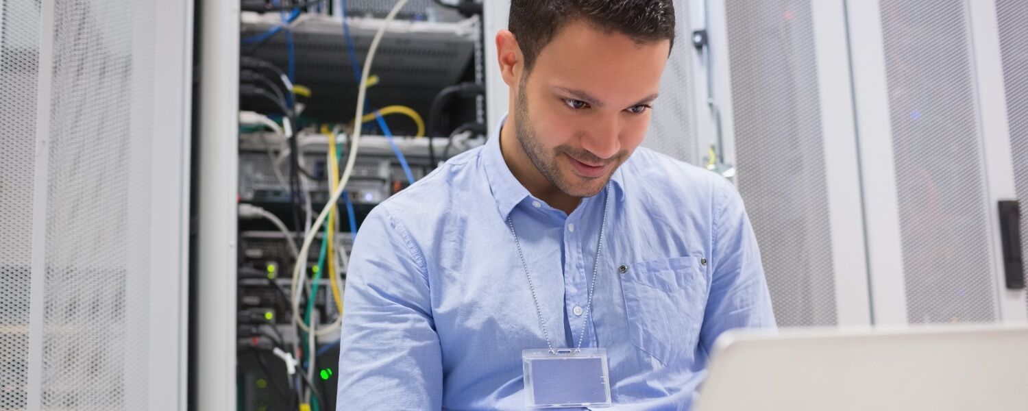 A man is sitting in front of a laptop computer in a server room.