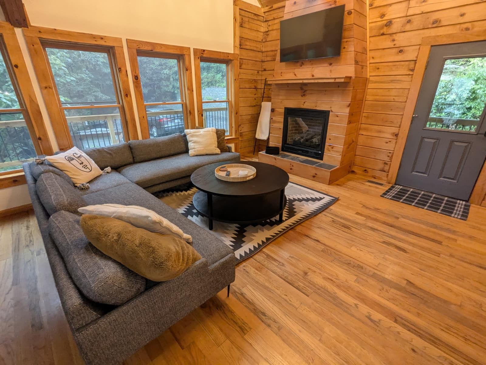 Cozy cabin living room with gray sectional sofa, fireplace, and large windows. Wooden walls and floors, natural light.