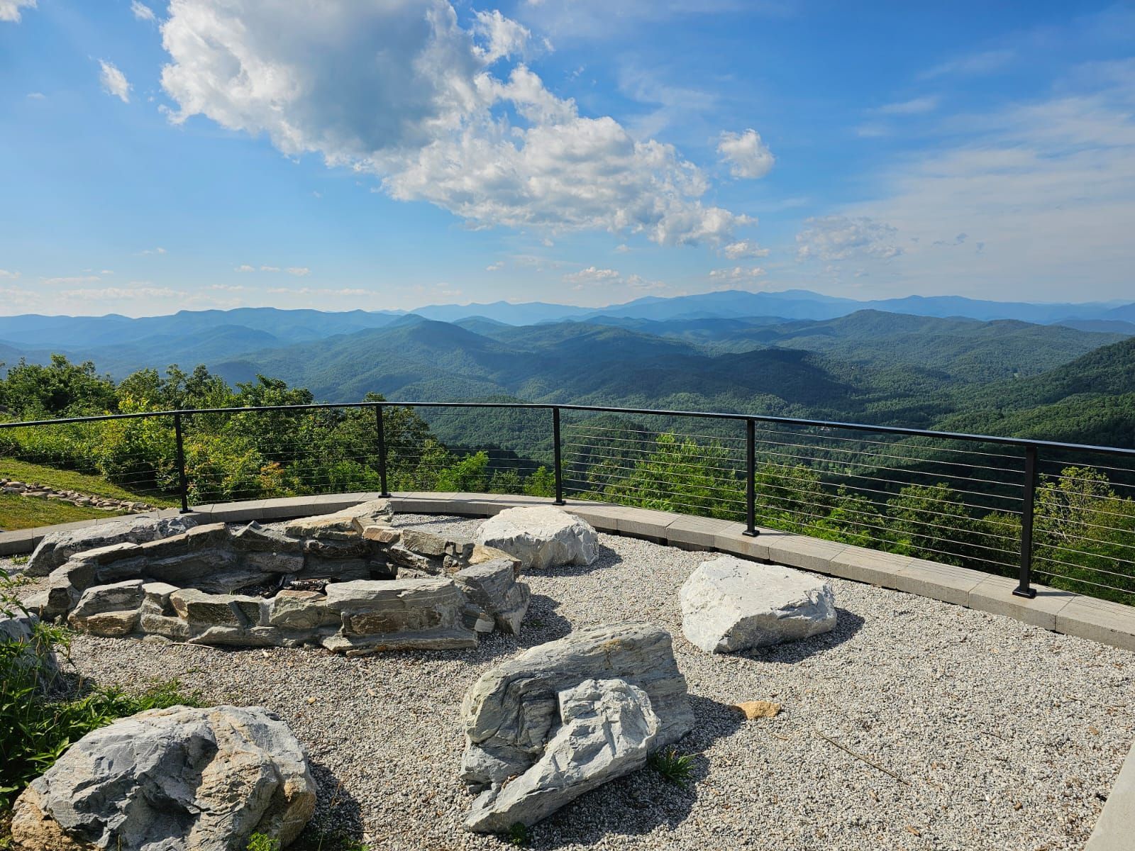 Fire pit on a mountain deck with a view of rolling green hills under a partly cloudy sky.