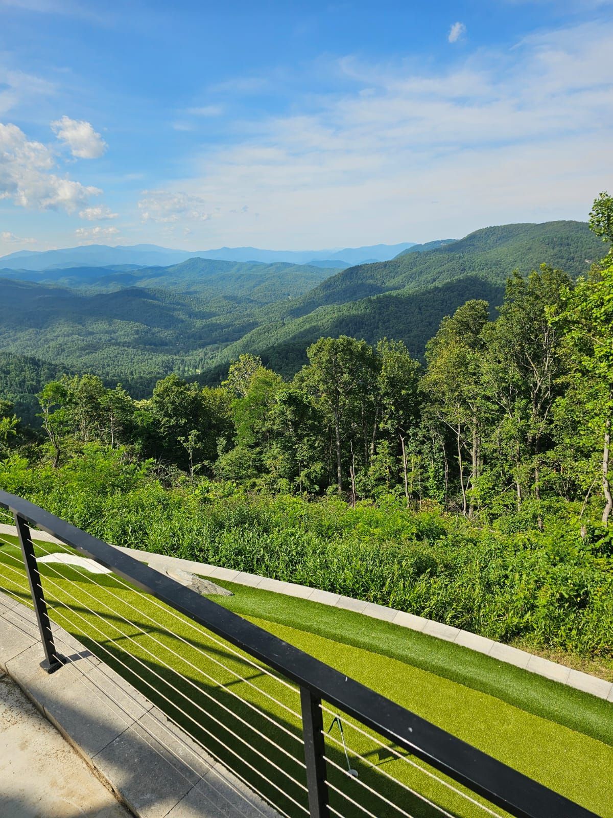 Mountain vista: lush green forests and rolling hills under a blue sky, viewed from a deck with a black railing.