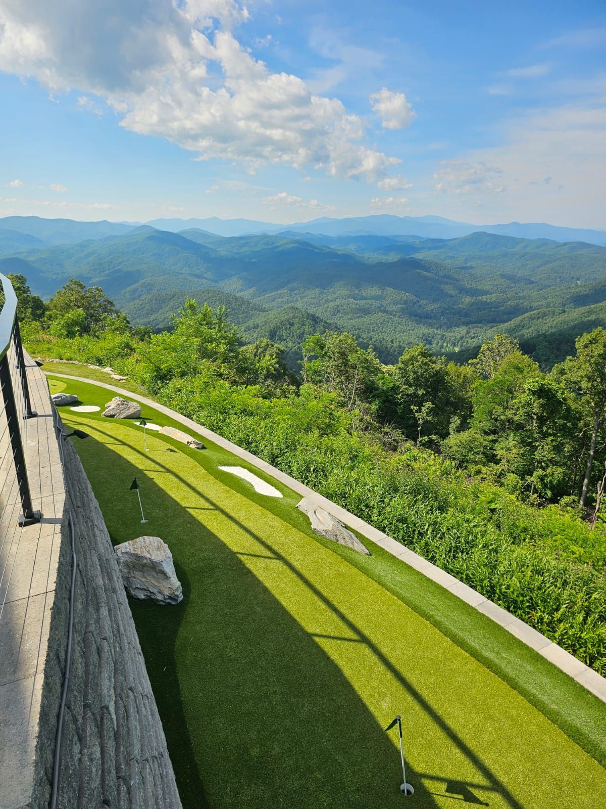 Mini golf course with mountain views. Green artificial turf, blue sky, trees, and distant hills.