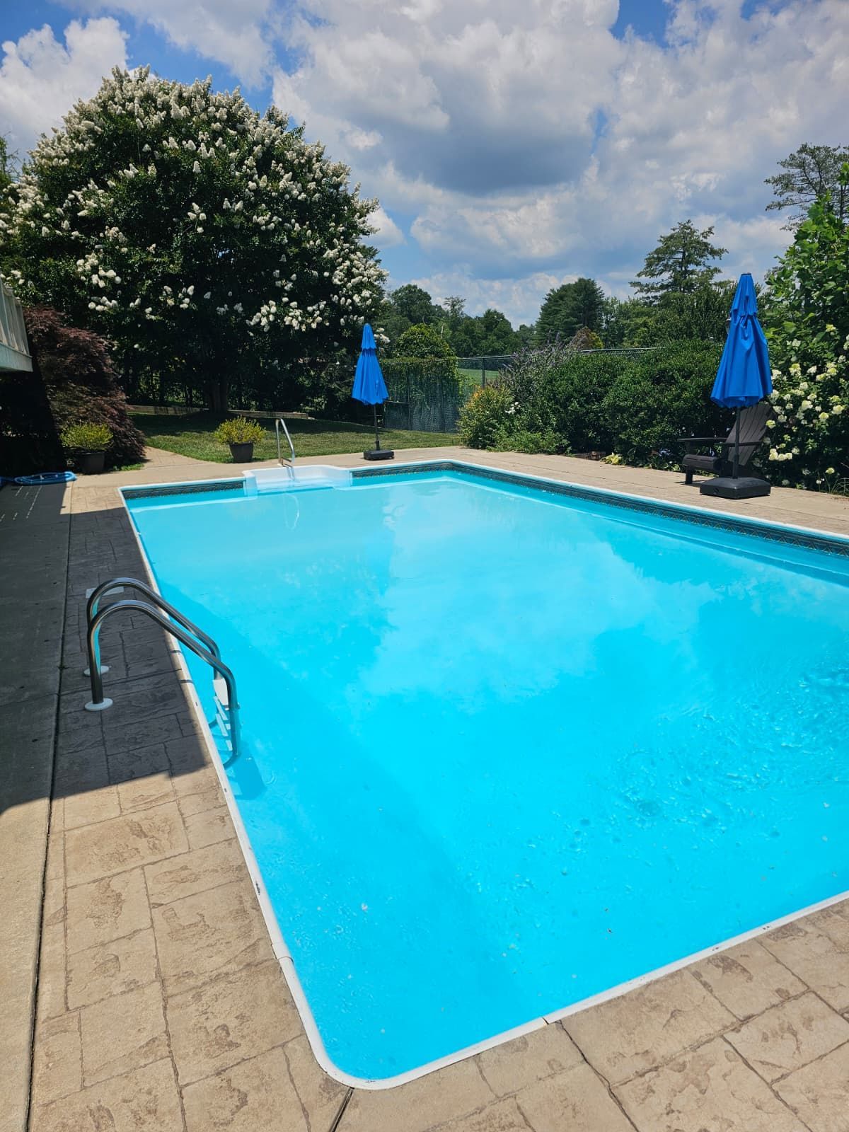 A rectangular outdoor swimming pool filled with bright blue water, flanked by patios and lush green trees, under a partly cloudy sky.