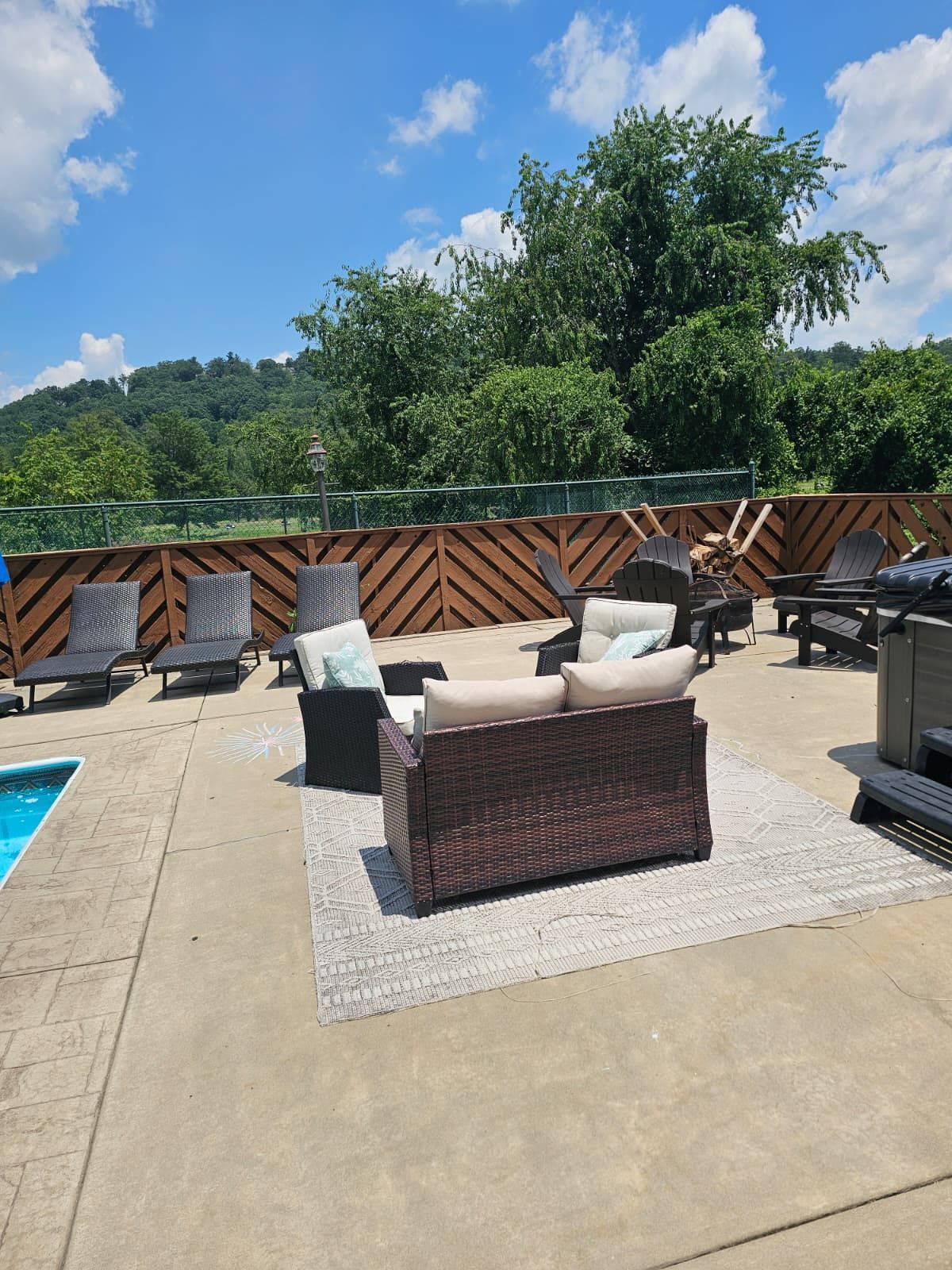 Outdoor patio with lounge seating by a pool, set against a wooden fence, with trees and a blue sky.