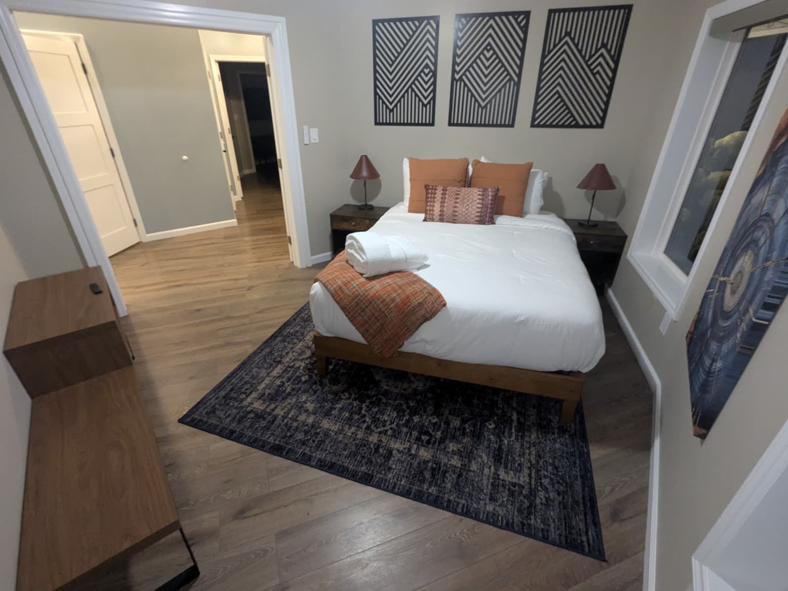 Bedroom with a bed, rug, art, and end tables. Wooden floor and walls are painted neutral colors.