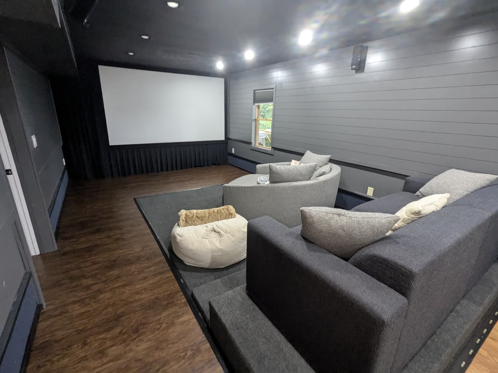 Home theater with a large screen, couches, and bean bag on a carpeted platform, dark grey walls, and wood flooring.