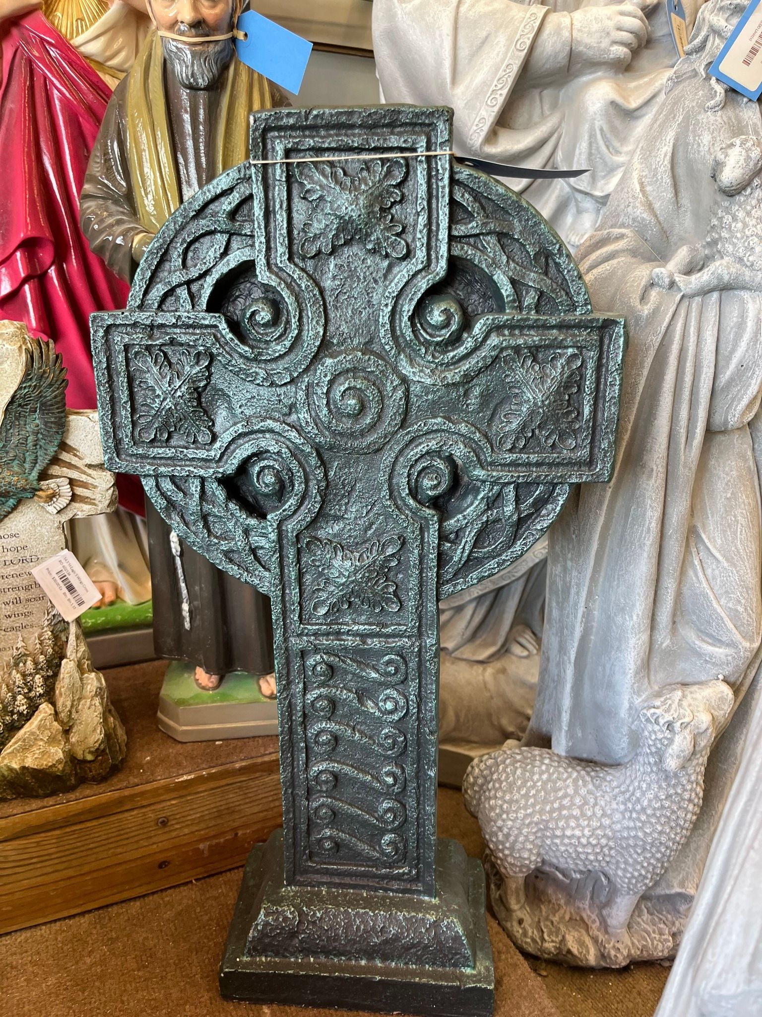 Celtic cross statue with intricate detailing, dark gray color, placed among other religious figures.