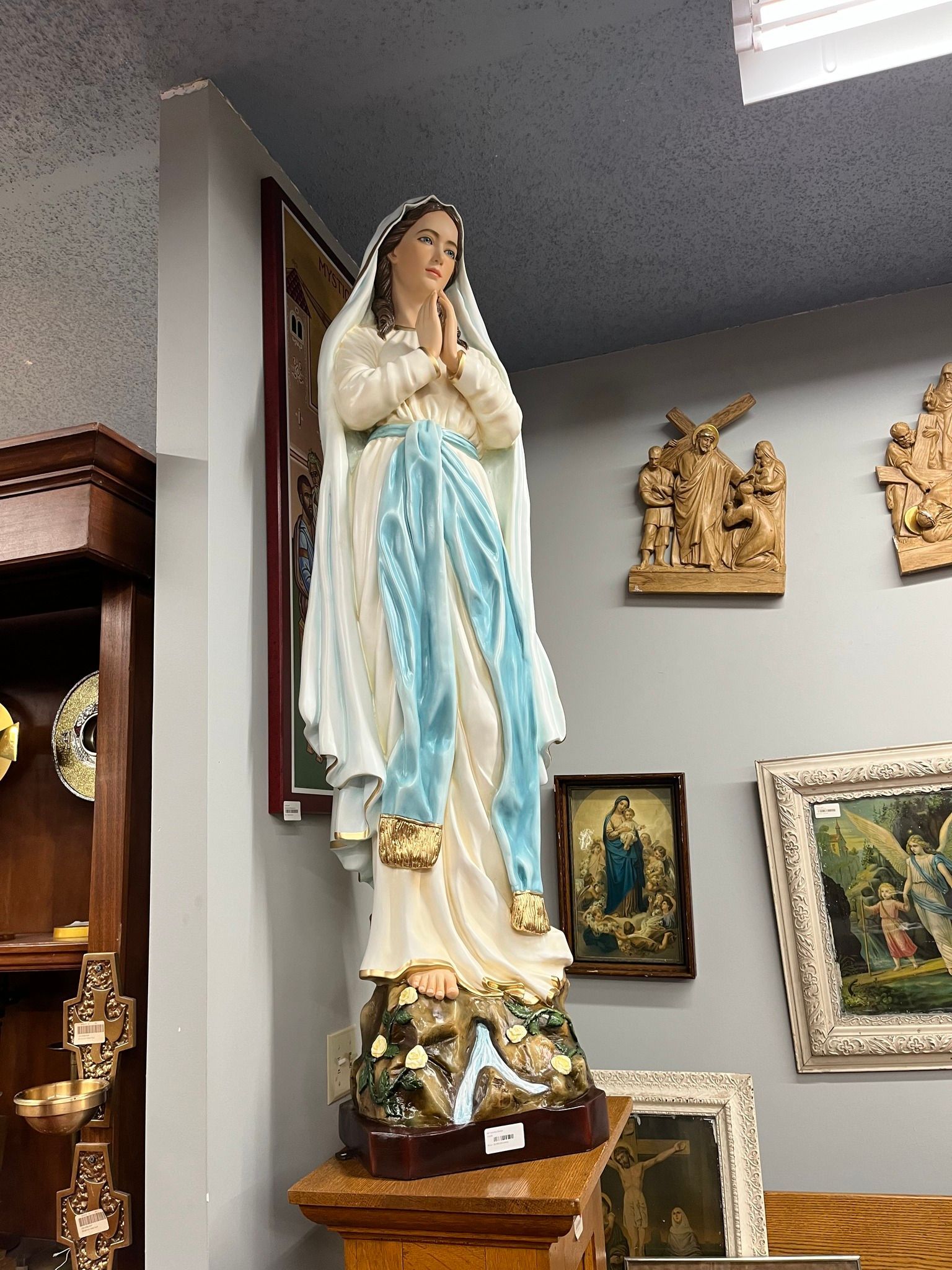 Statue of the Virgin Mary with blue and white robes, hands clasped in prayer, in a room with other religious items.