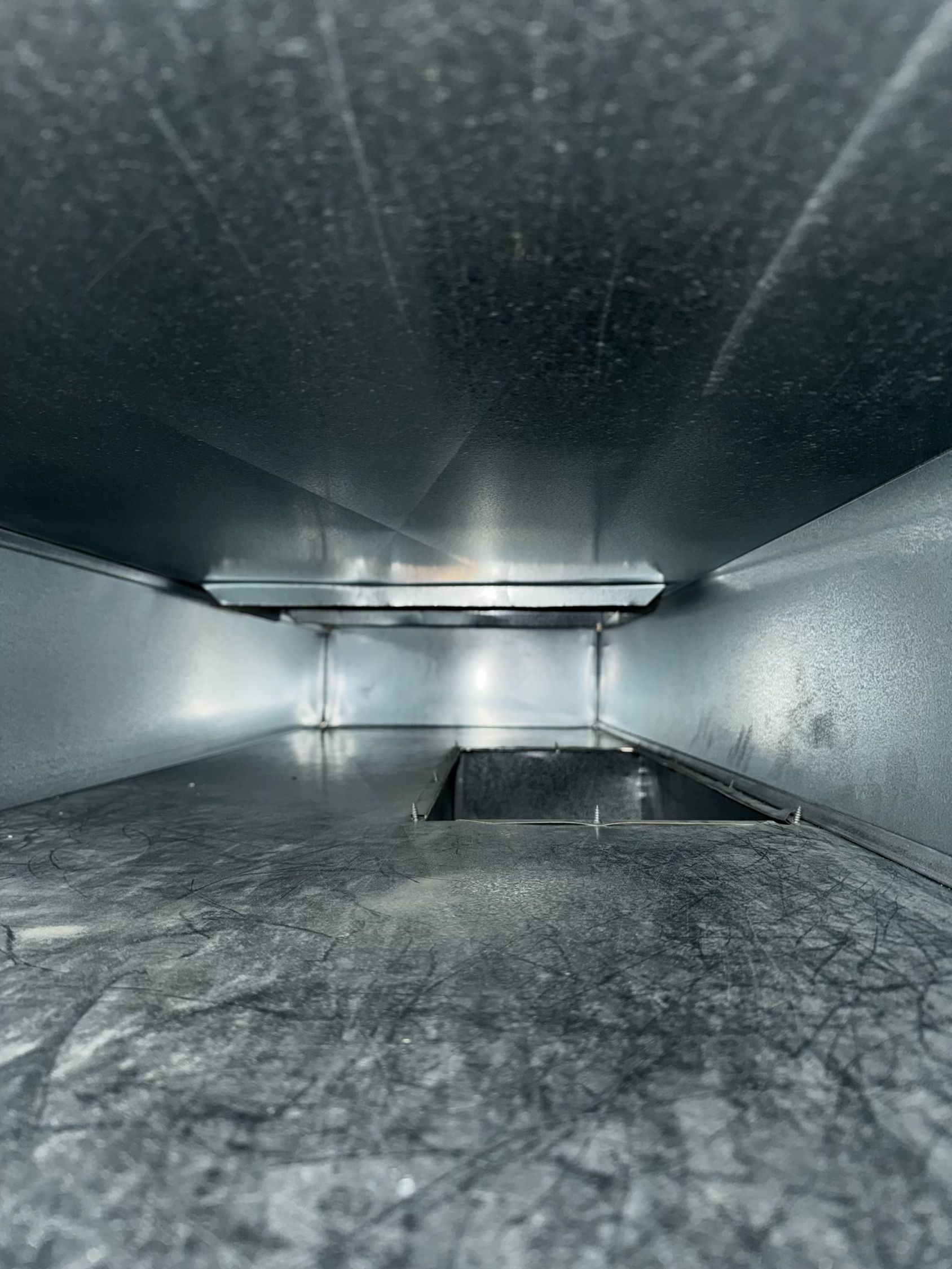 Inside view of a rectangular HVAC duct, silver-colored metal with a small opening and debris visible on the bottom.