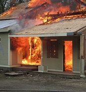 House engulfed in flames, fire bursting from the porch and roof, smoke billowing upwards.