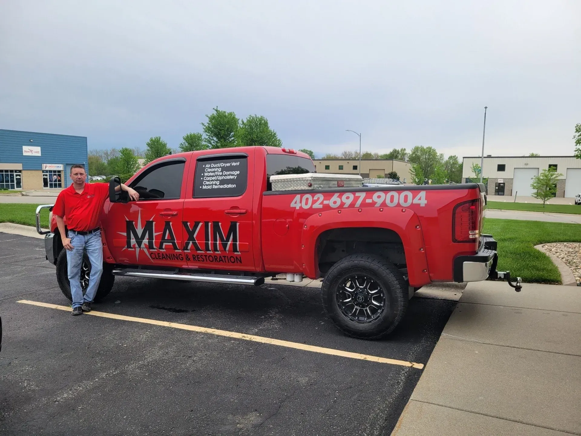 Man stands next to red truck with Maxim Painting & Interior logo. Phone number 402-697-9004. Parked outside buildings.