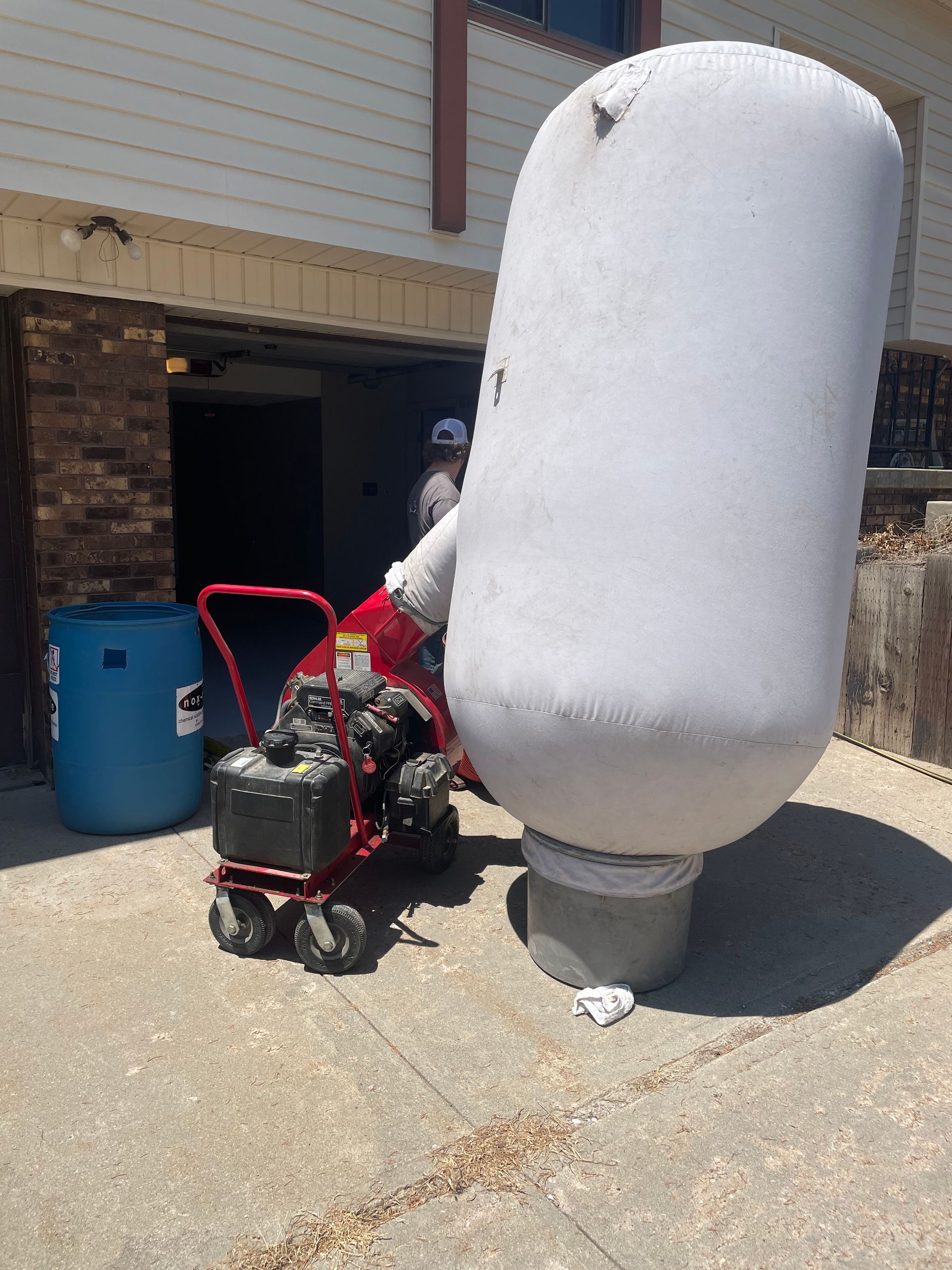Red cart with machine, large white container, blue barrel by building. Person in background.