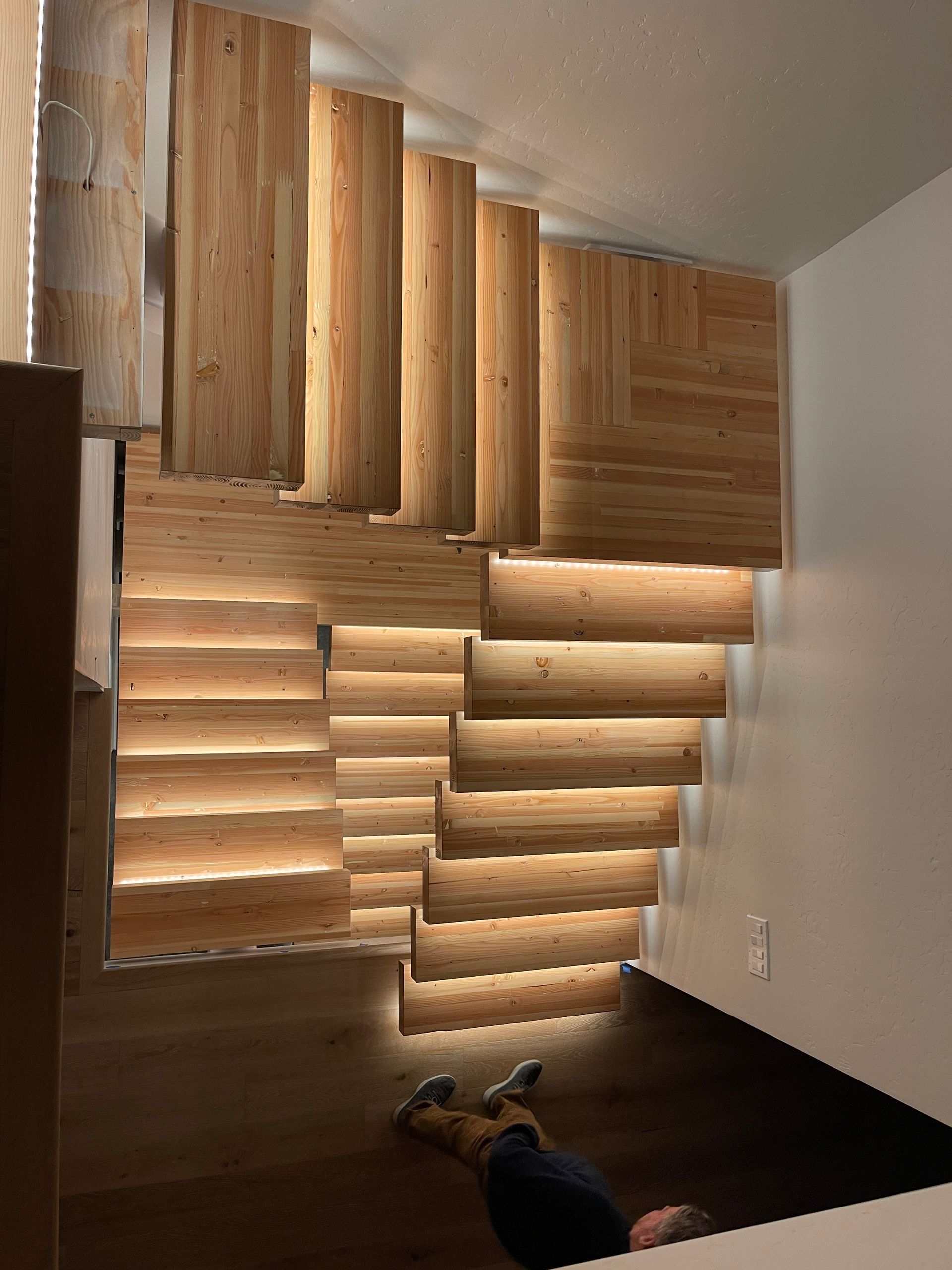 A person is laying on the floor under a wooden staircase.