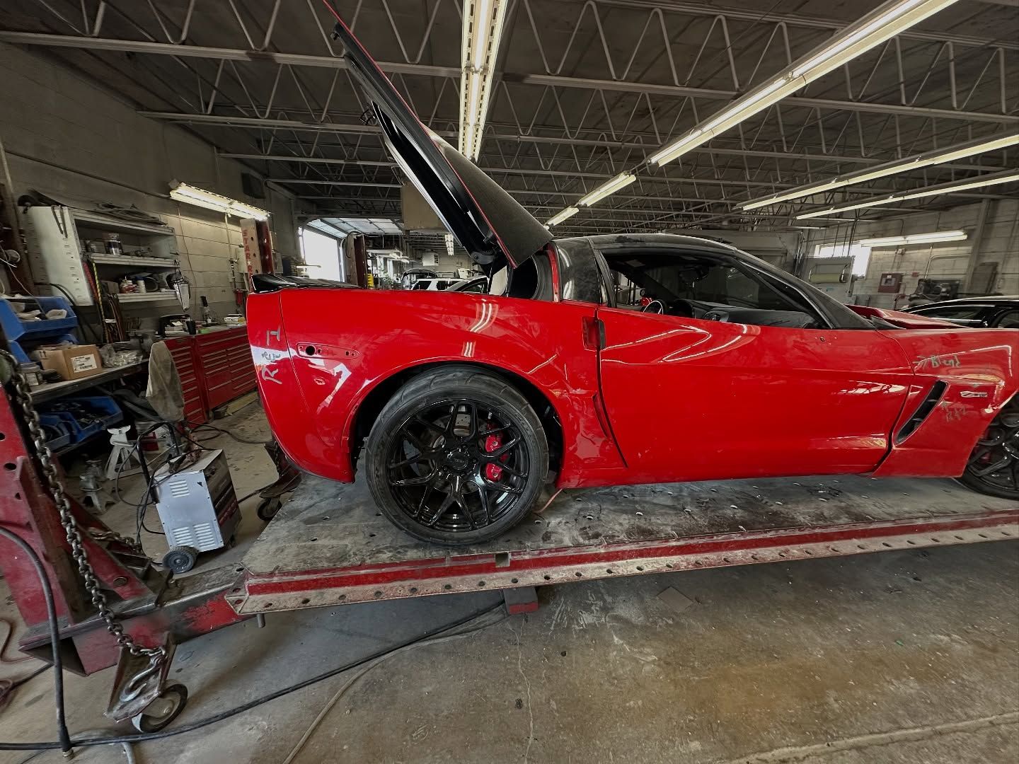 Red Corvette with black wheels, damage on the rear panel, parked on a tow truck in a repair shop.