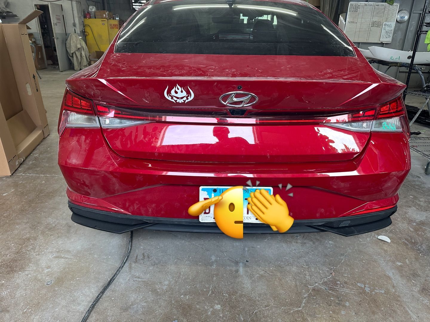 Red car's rear, with a license plate and stickers. A yellow emoji covers the plate. Indoors.