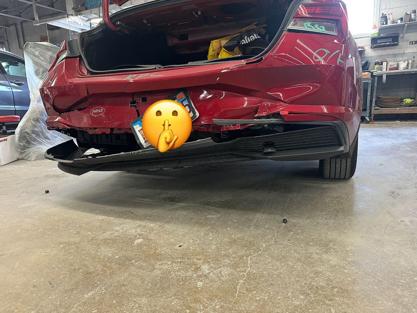 Red car with rear-end damage at a repair shop; bumper torn off.