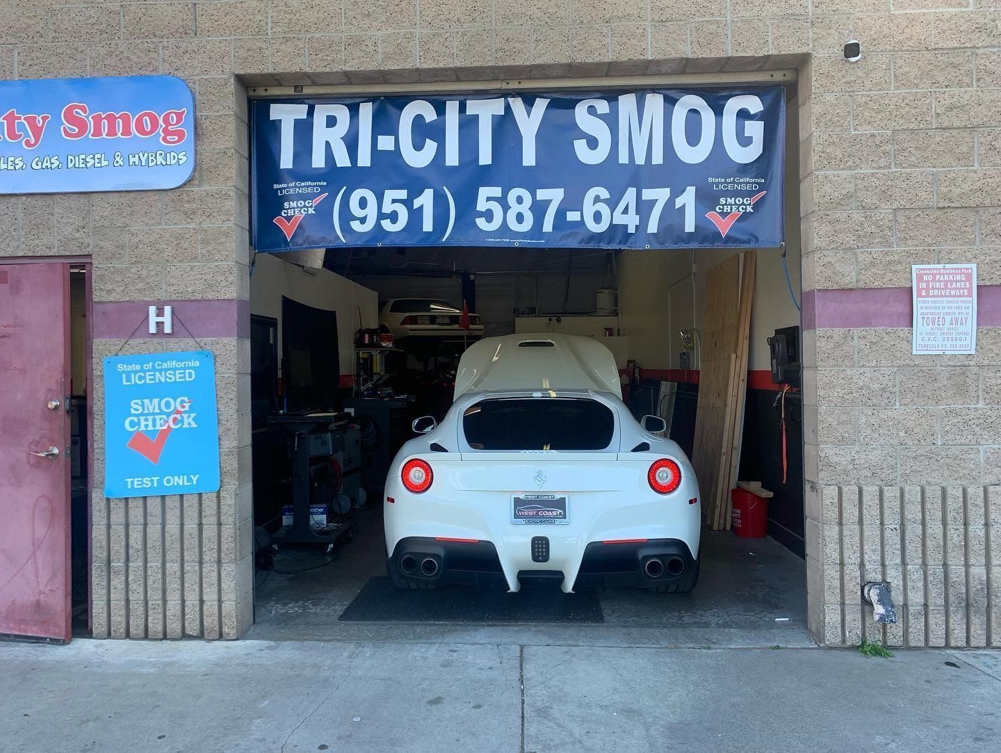 A white Ferrari inside the bay of a smog check station called Tri-City Smog in front of a blue sign with a phone number.