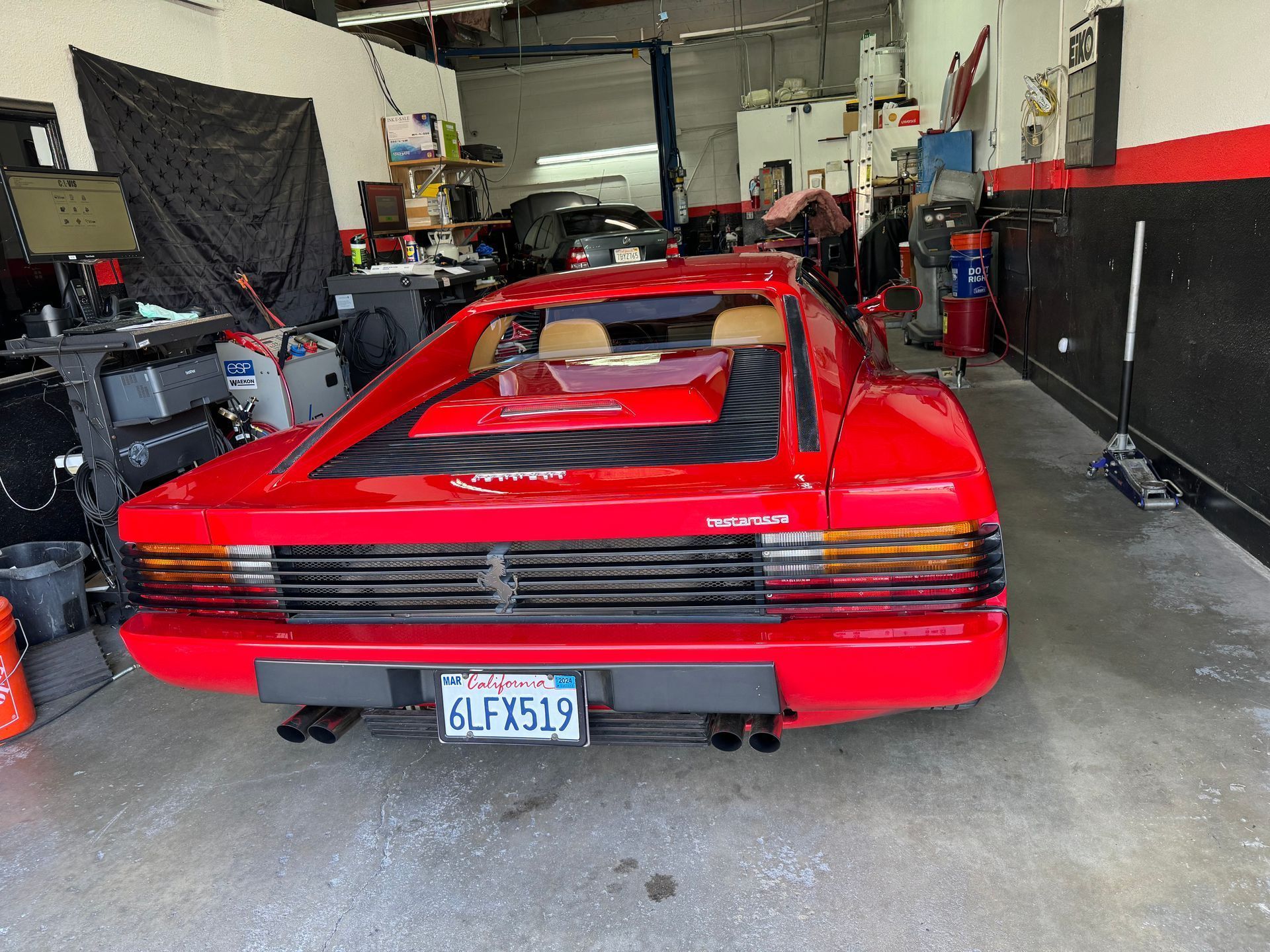 Red Ferrari Testarossa parked inside a car repair shop. The license plate is visible and the shop appears cluttered.