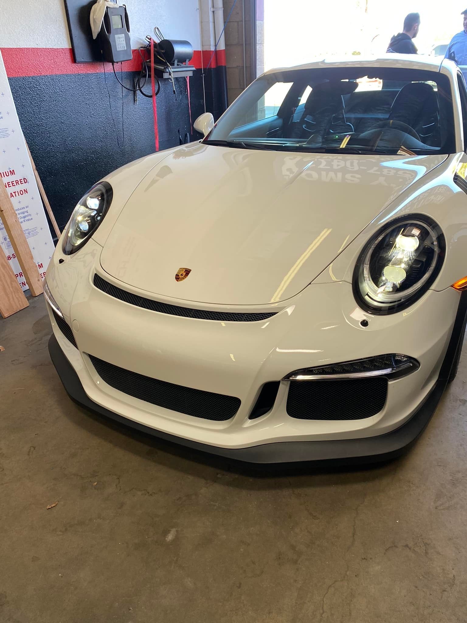 White Porsche 911 GT3 in a garage. The car's front is visible, showcasing its headlights, front bumper, and the Porsche emblem.