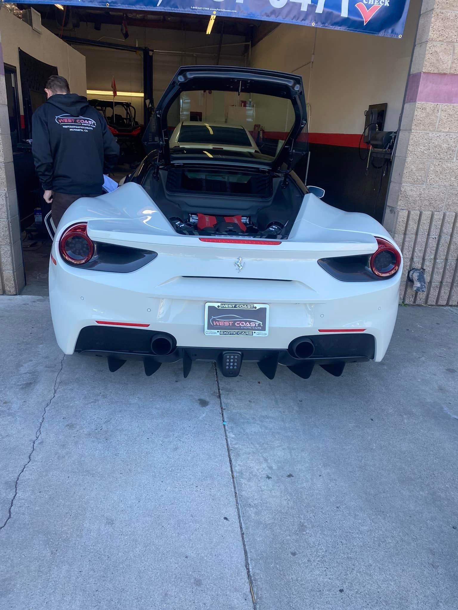White Ferrari 488 Spider with its trunk open, parked inside a garage. A person stands nearby.
