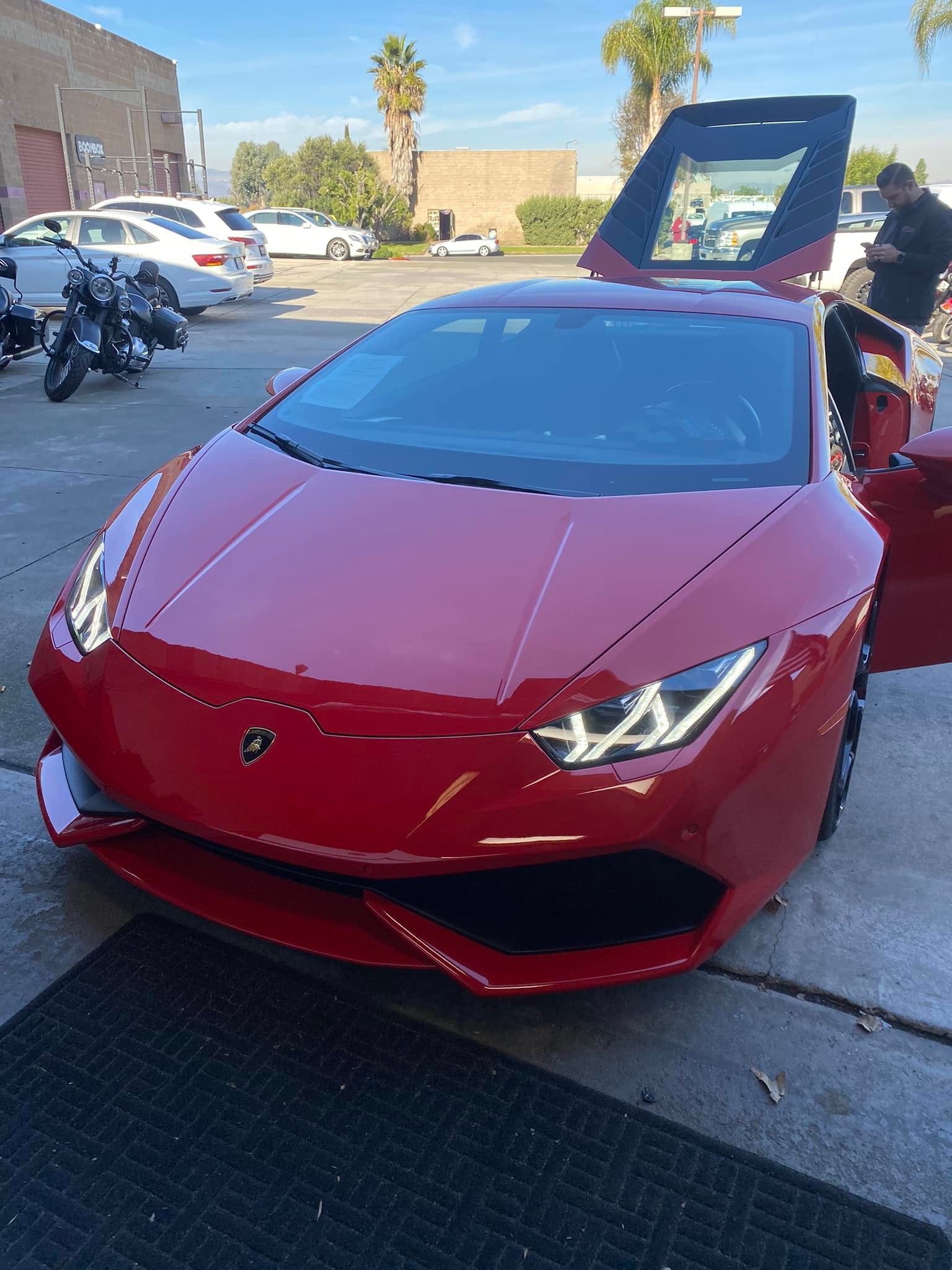 Red Lamborghini with doors open, parked outside on a sunny day. A person stands near the car.
