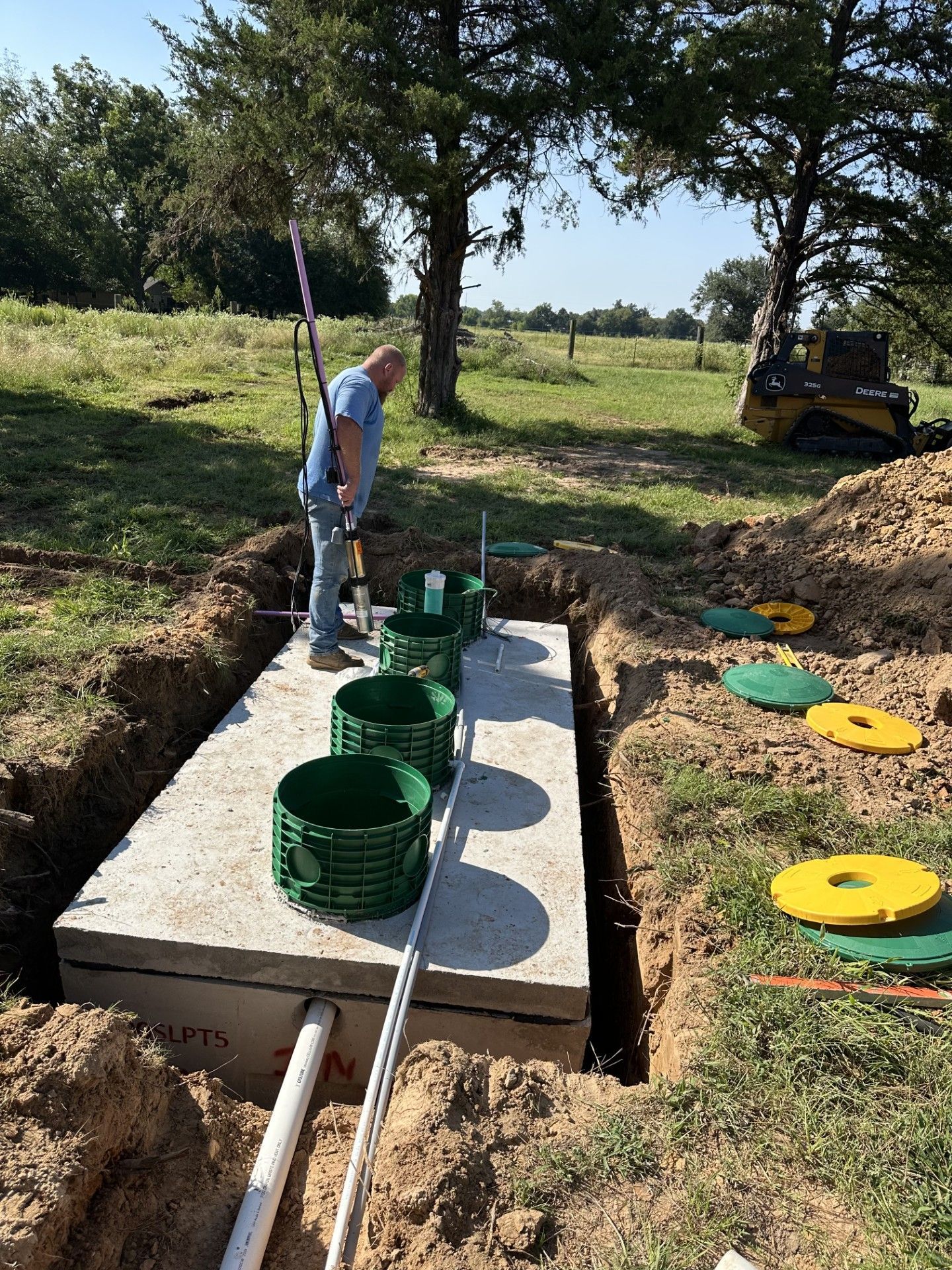 Yellow excavator lifting a concrete septic tank into a hole in a wooded area.