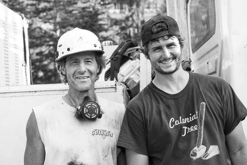 Two workers in hard hats smiling outdoors, one wearing a respirator around his neck.