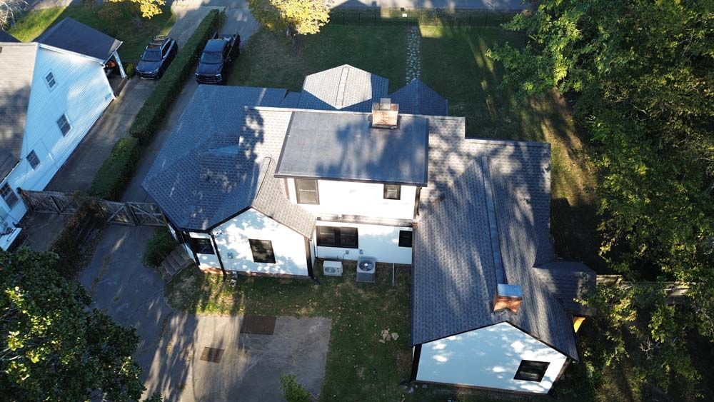 Aerial view of a white house with dark gray roof, surrounded by trees and a driveway with two cars.