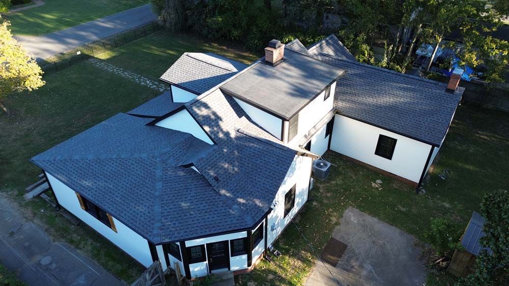 Aerial view of a white house with black trim and a dark gray roof, surrounded by green grass and trees.