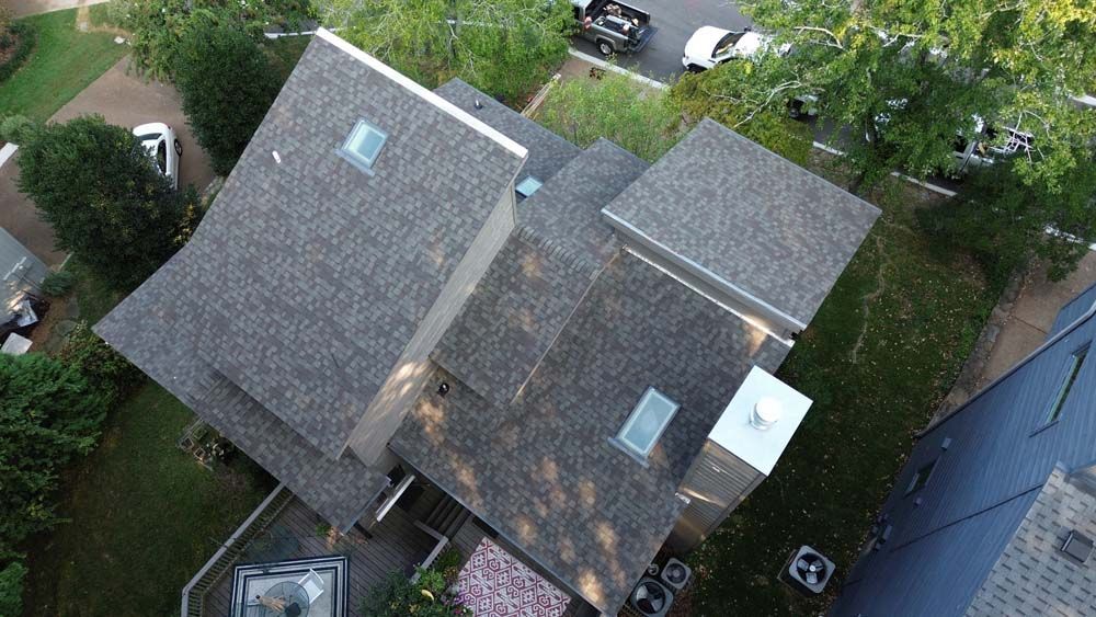 Aerial view of a multi-level house with a gray shingle roof, skylights, and a white chimney.