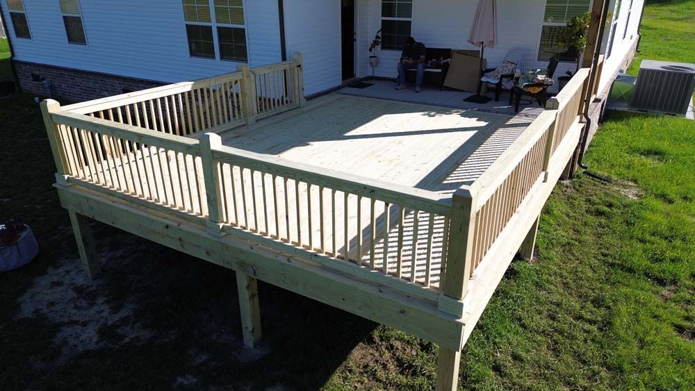 Wooden deck attached to a white house with railing and supports, on a grassy lawn.