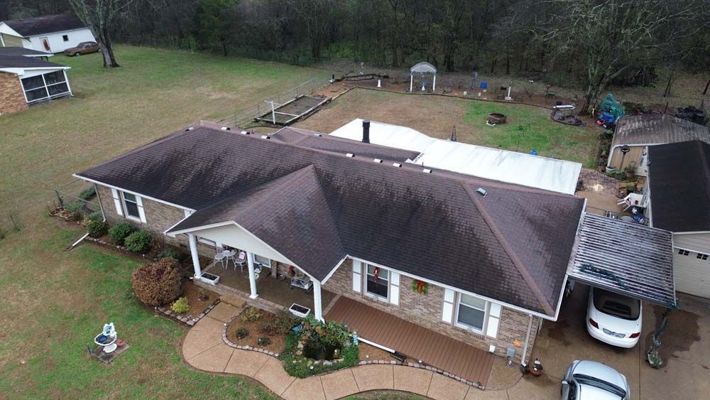 Drone view of a one-story brick house with a dark roof and a carport, surrounded by a yard.