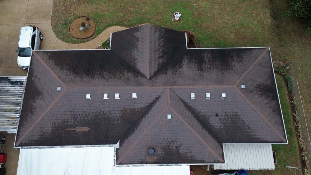 Aerial view of a brown asphalt shingle roof on a house, with white trim and a few vents, and a white van parked nearby.