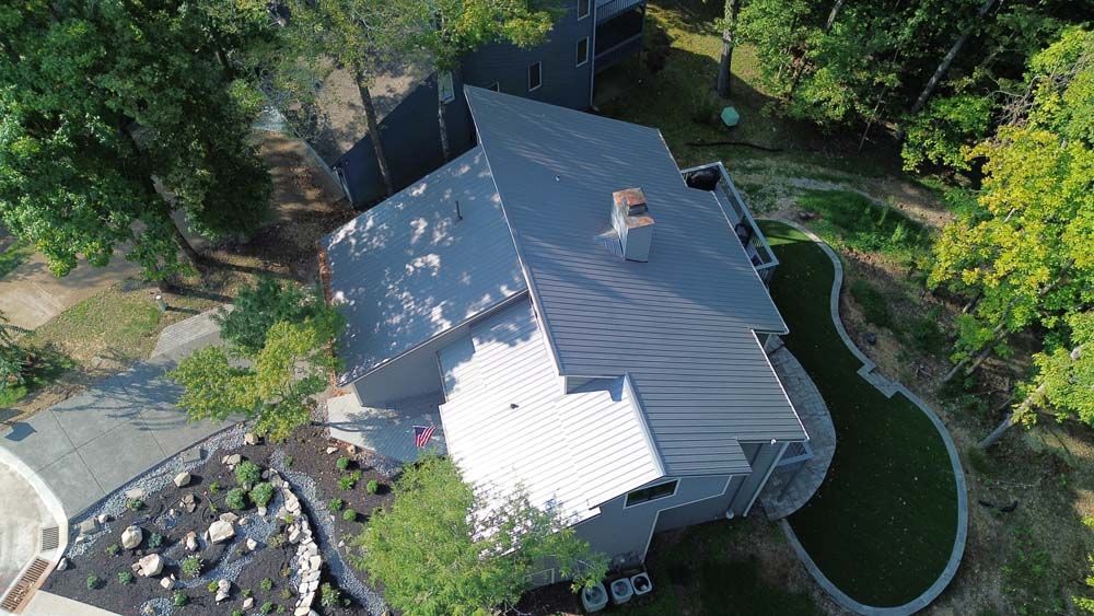Aerial view of a modern gray house with a metal roof surrounded by trees and a circular driveway.