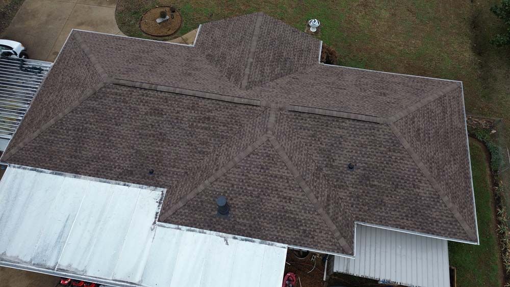 Aerial view of a brown asphalt shingle roof on a house, with white siding and green yard.