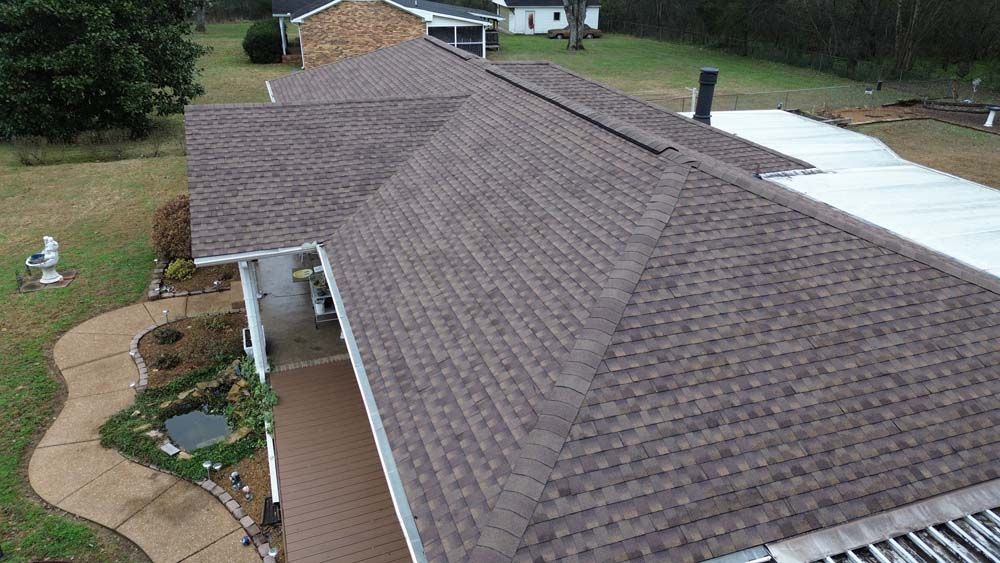 Brown shingled roof of a house with a porch and surrounding yard.