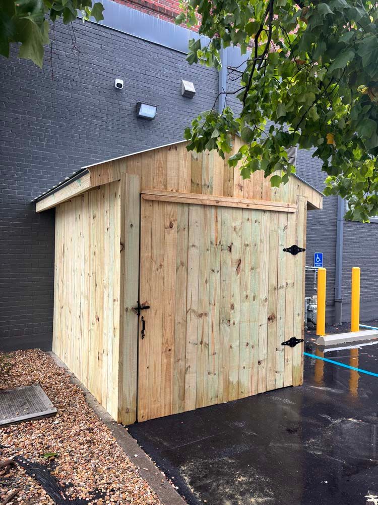 Wooden storage shed next to a dark brick building. Black hardware, asphalt, and blue curb.