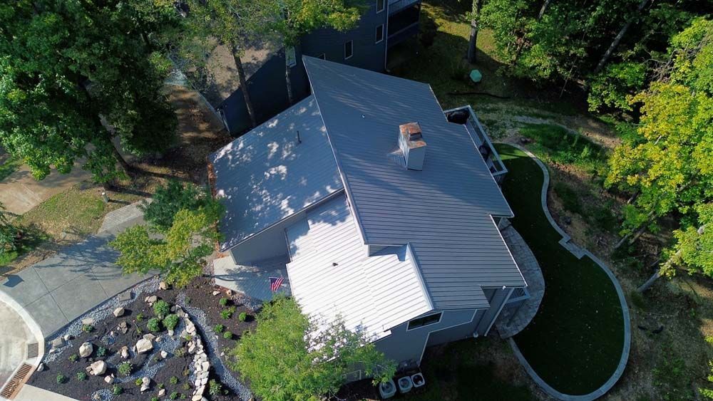 An aerial view of a modern house with a metal roof surrounded by trees and a curving lawn.