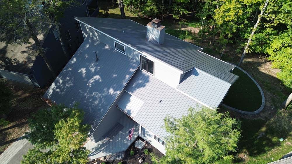 Aerial view of a modern house with a gray metal roof, chimney, and surrounding trees.