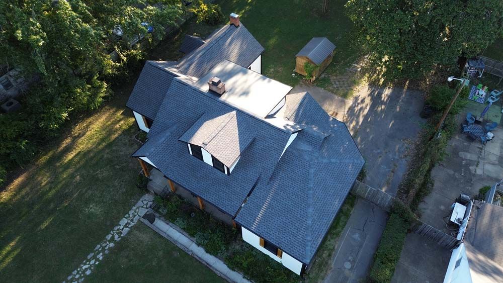 Overhead view of a house with a dark gray roof, dormers, chimney, and a driveway in a green yard.