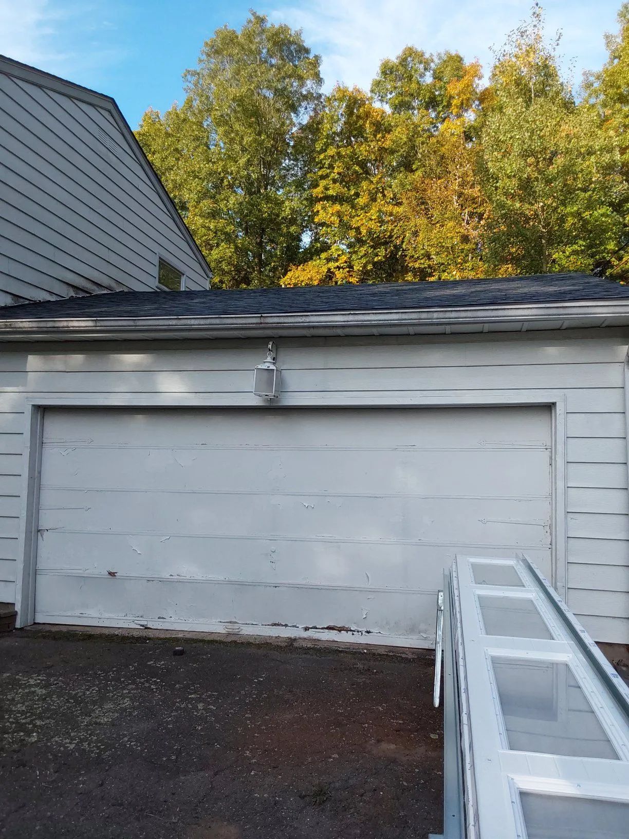 White garage door beside a house, with dark roof shingles and autumn trees in the background.