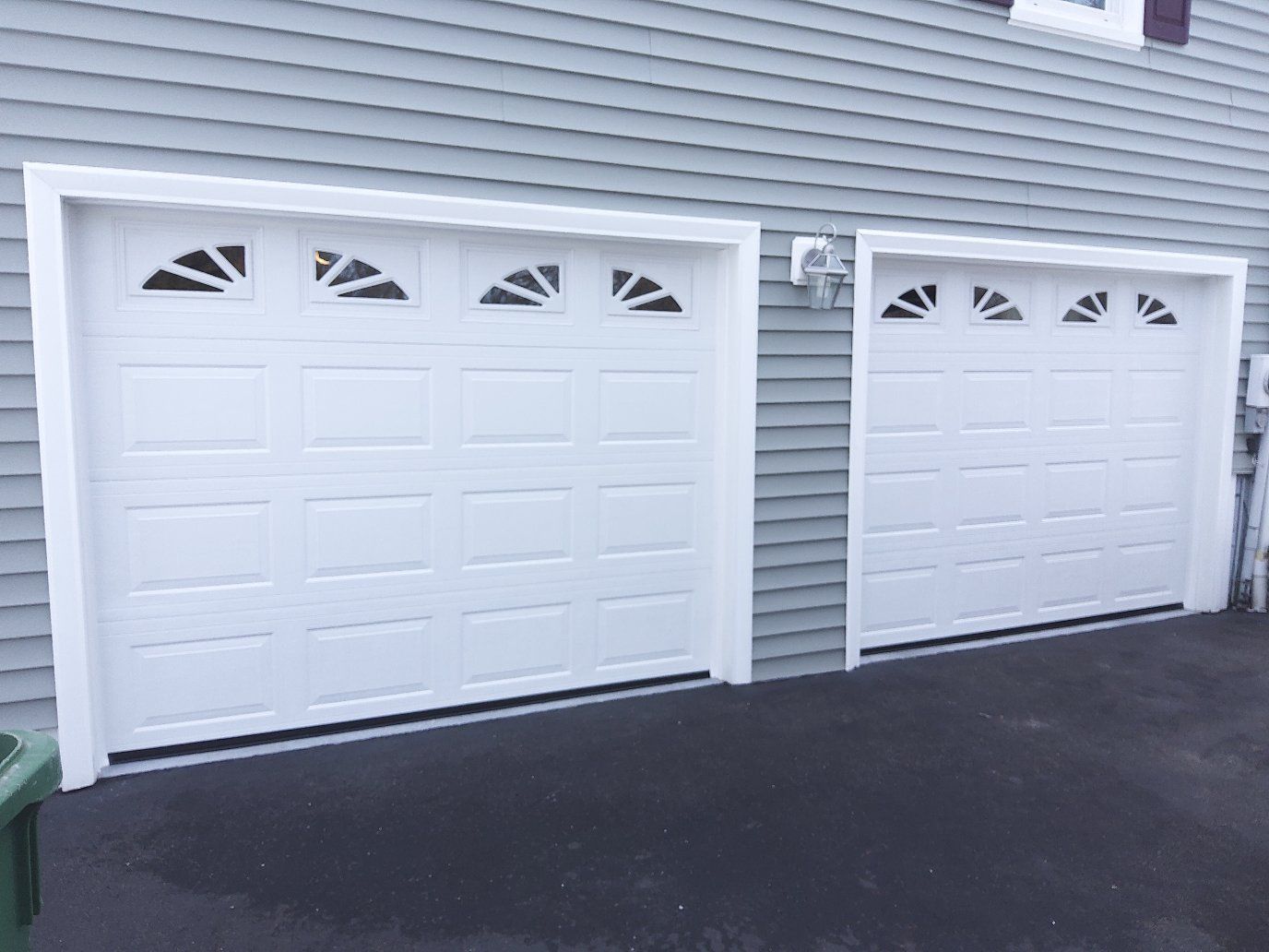 Two white garage doors with decorative windows on a gray house exterior and dark driveway
