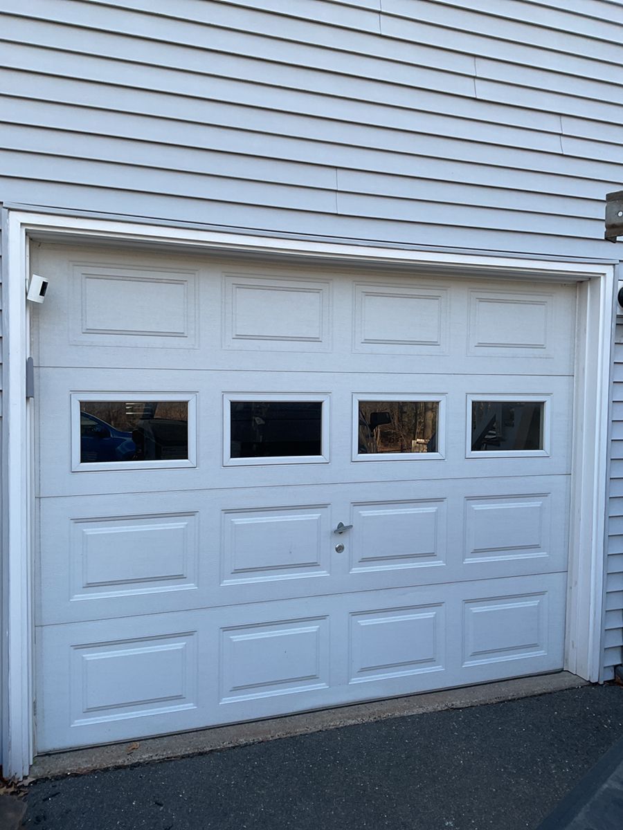 White garage door with four small windows, set in light siding beside a driveway