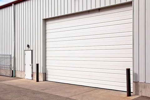 Large white roll-up garage door on a gray industrial building with a small side door and bollards