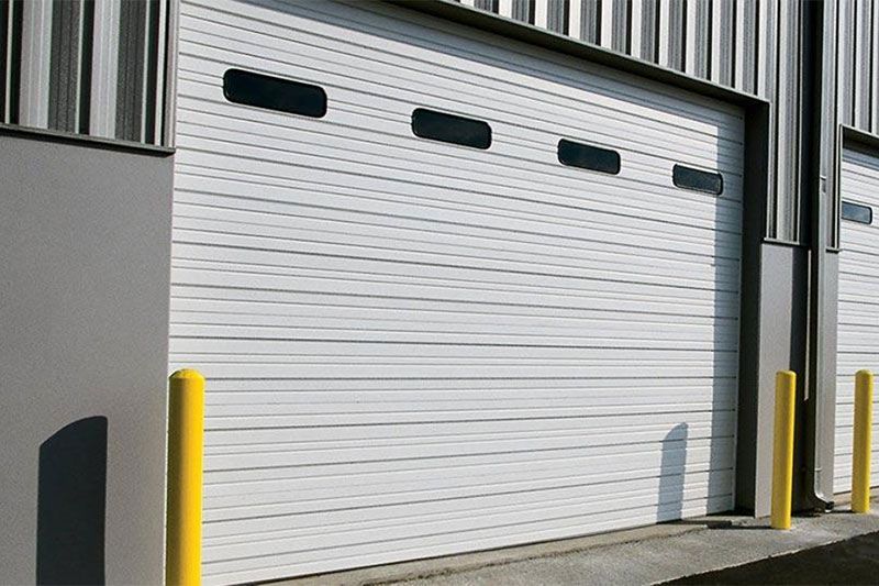 White industrial roll-up garage door with small windows, flanked by yellow safety bollards.