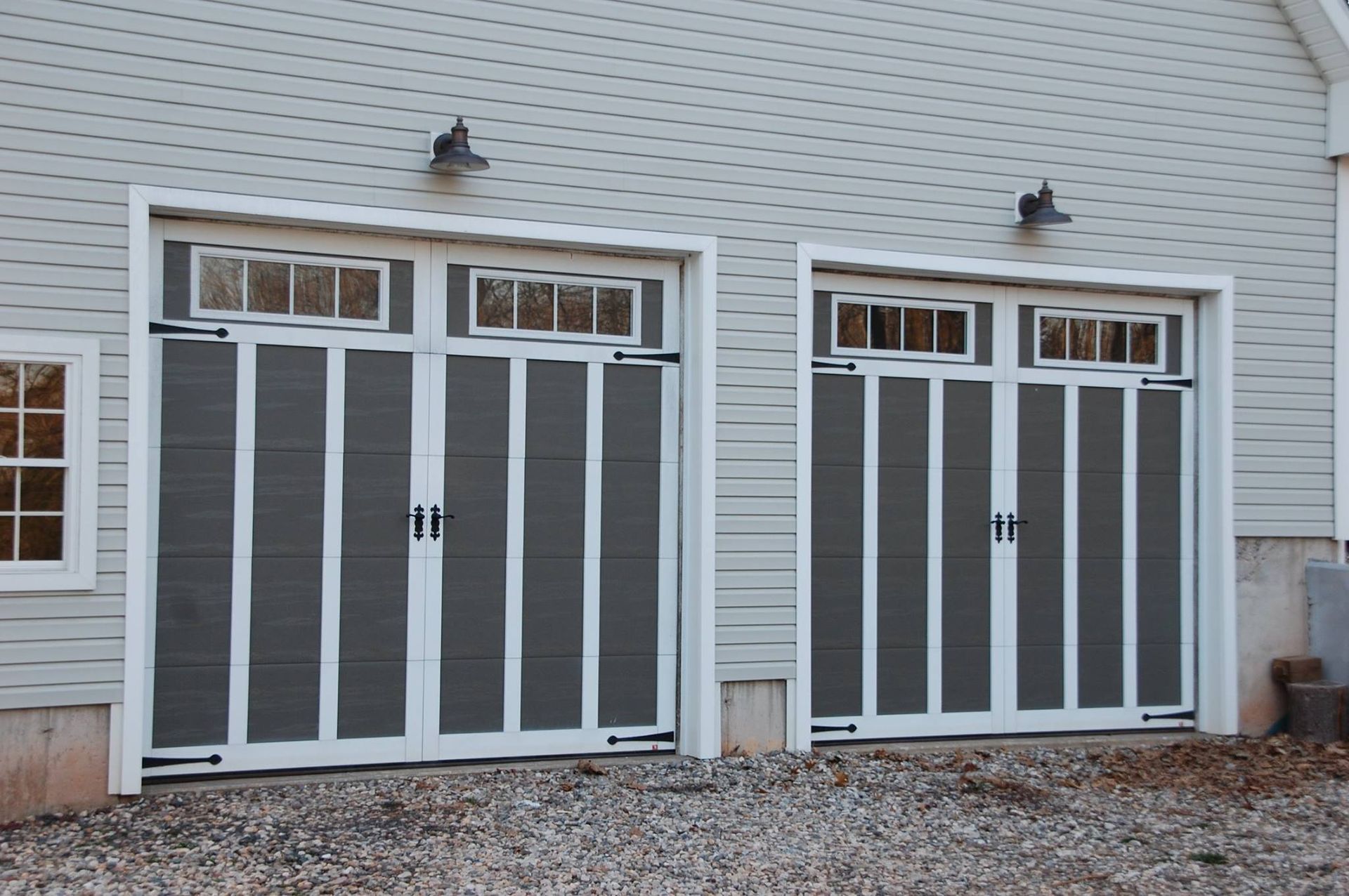 Two white-framed garage doors on a gray house exterior with small windows and wall-mounted lights