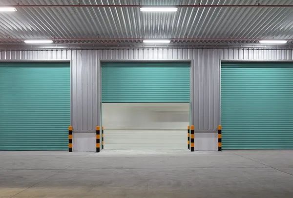 Three teal warehouse loading bay doors on a gray industrial wall with yellow-black safety bollards