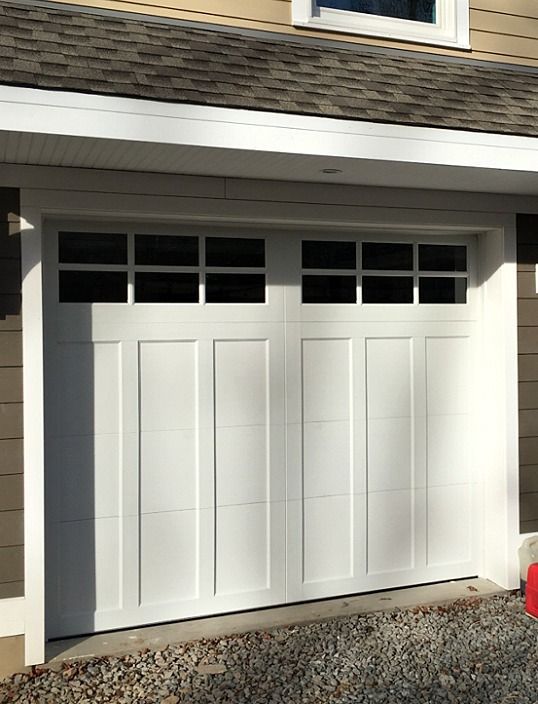 White paneled garage door with small windows under a house overhang, beside a gravel driveway.