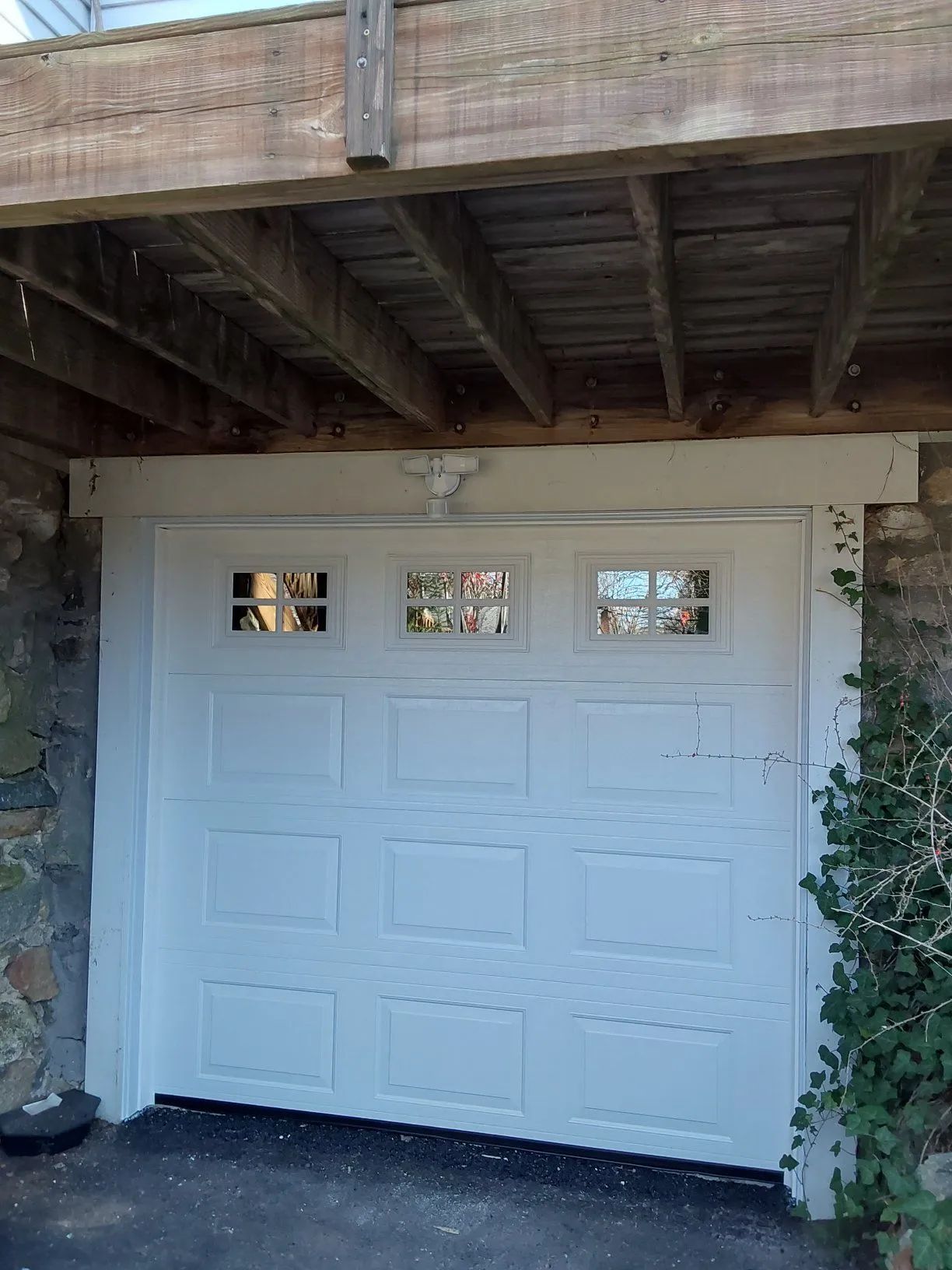 White garage door beneath a wooden deck, framed by stone walls and ivy.
